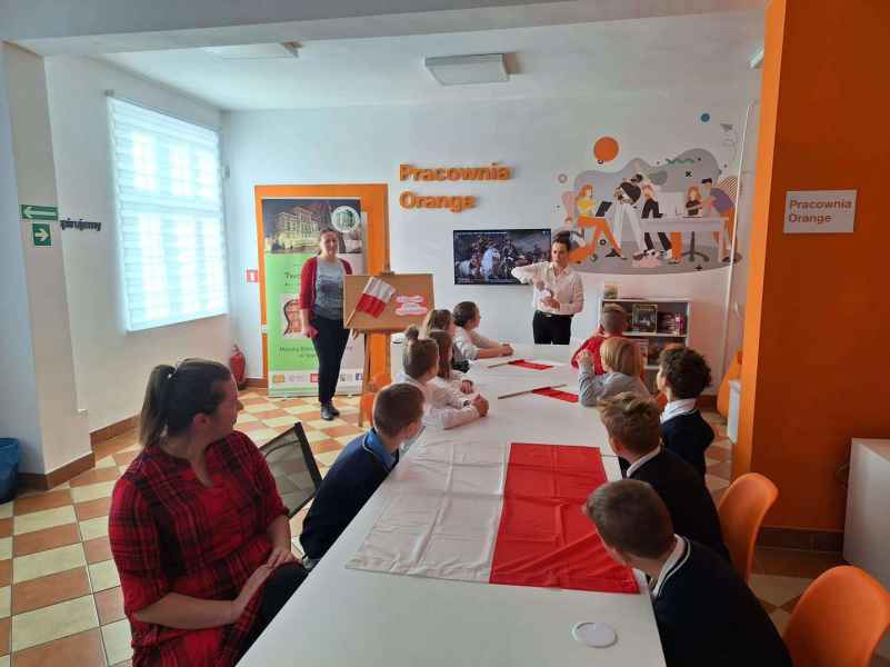 zdjęcie bibliotekarki pokazującej dzieciom siedzącym przy stoliku z flagami Polski kotylion