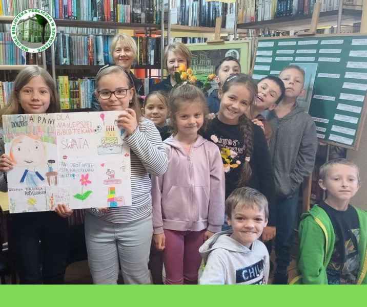 grupowe zdjęcie bibliotekarki z kwiatami, dzieci i wychowawczyni z plakatem z życzeniami, po lewej stronie logo biblioteki