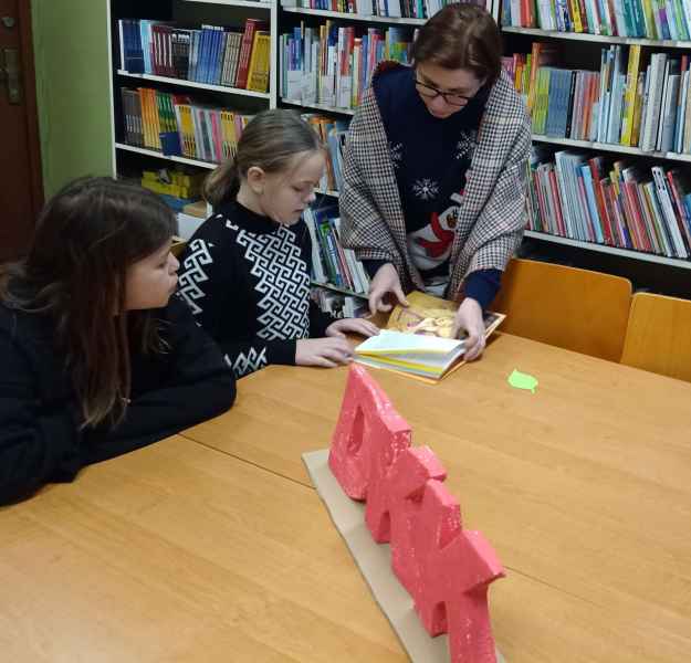 zdjęcie bibliotekarki pokazującej dzieciom miejsce czytania w książce, na stole logo DKK ze styropianu