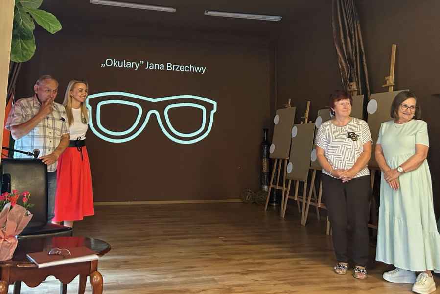 zdjęcie dyrektor biblioteki oraz seniorów na scenie, w tle slajd prezentacji z napisem "okulary" - Jana Brzechwy