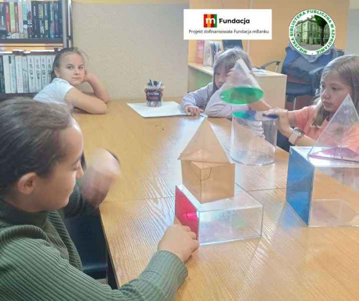 zdjęcie dzieci przy stole podczas oglądania brył geometrycznych, na górze logo biblioteki i mfundacji z napisem projekt dofinansowała fundacja mbanku
