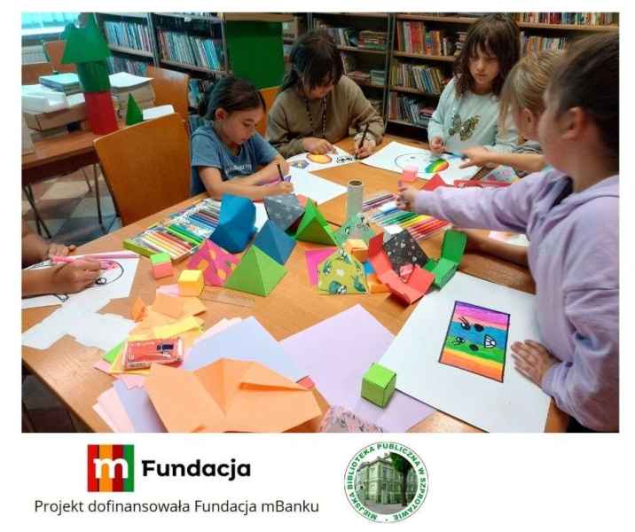 zdjęcie dzieci przy stoliku podczas kolorowania rysunków figur geometrycznych, na dole logo mfundacji z napisem projekt dofinansowała fundacja mbanku i logo biblioteki