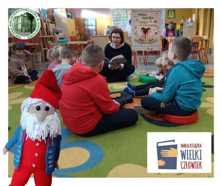 zdjęcie bibliotekarki podczas czytania dzieciom książki na dywanie, w górze logo biblioteki, na dole logo projektu mała książka wielki człowiek i dziergana maskotka krasnala