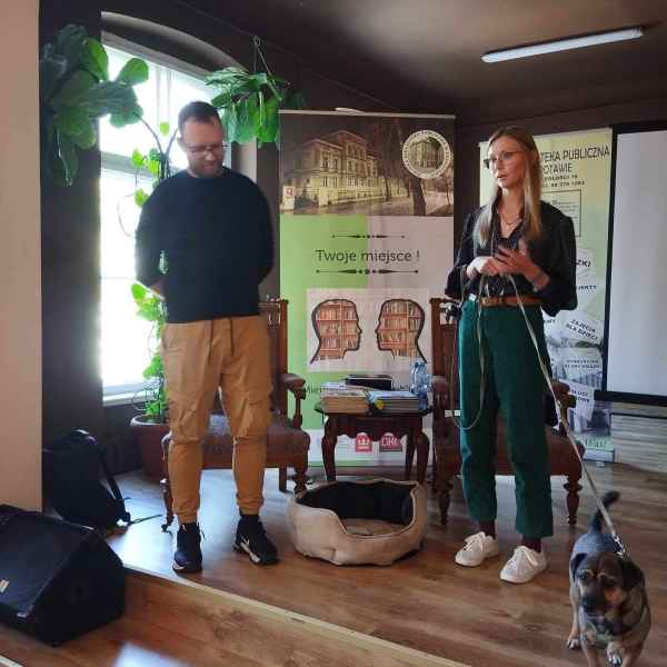 zdjęcie mężczyzny oraz autorki trzymającej psa na smyczy na tle baneru biblioteki