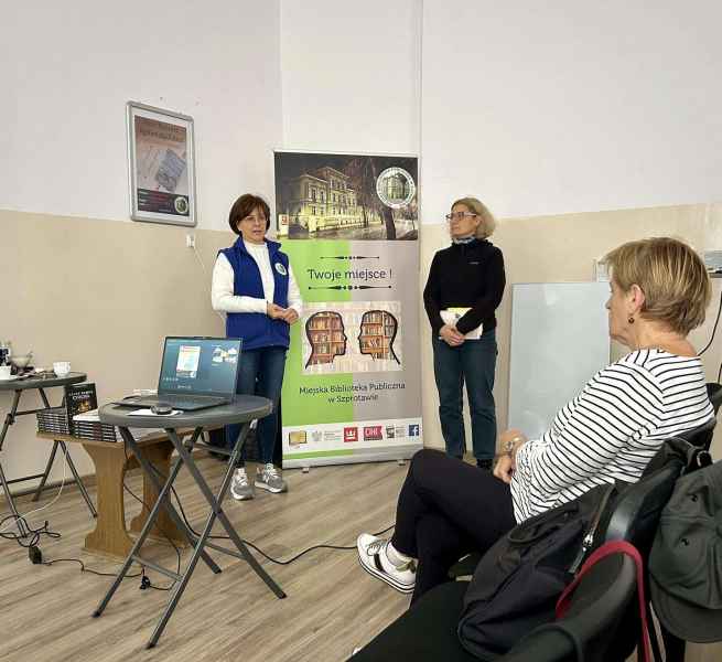 zdjęcie bibliotekarki, pisarki oraz pierwszego rzędu czytelników, z tyłu baner biblioteki