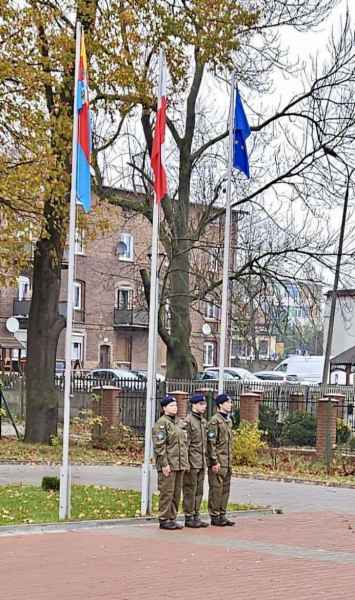 zdjęcie trojga uczniów w strojach wojskowych stojących pod flagą Polski