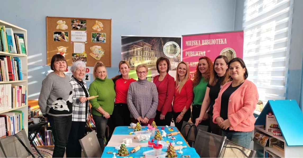 zdjęcie dyrektor biblioteki, bibliotekarki oraz uczestników klubu DKK przy stoliku z piernikami, w tle baner biblioteki i DKK