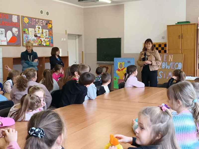 zdjęcie dzieci siedzących przy stolikach i nauczycielek mówiących do nich