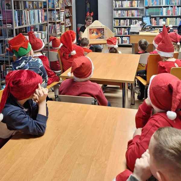 zdjęcie dzieci w czapkach Mikołaja i czerwonych strojach podczas oglądania teatrzyku kamishibai prezentowanego przez bibliotekarkę 