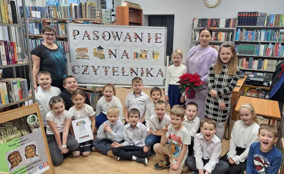 grupowe zdjęcie bibliotekarki, dzieci oraz wychowawczyni przy tablicy z napisem pasowanie na czytelnika