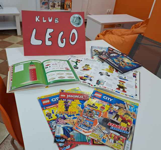 zdjęcie stolika z rozłożoną prasą i książkami o klockach lego, w tle plakat z napisem klub lego