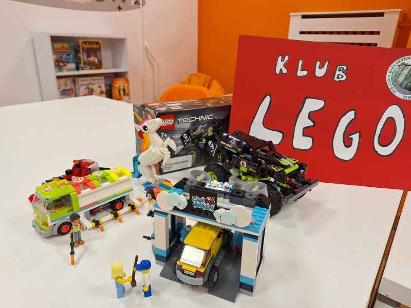 zdjęcie ułożonych modeli śmieciarki, auta terenowego i myjni samochodowej z klocków lego, z tyłu plakat z napisem klub lego i logo biblioteki