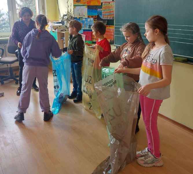 zdjęcie dzieci trzymających worki do segregacji odpadów i bibliotekarki instruującej dziecko gdzie wrzucać przykładowe opakowania