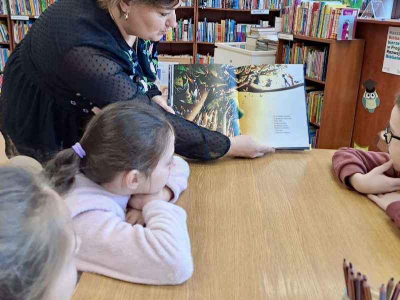 zdjęcie bibliotekarki pokazującej dzieciom przy stoliku książkę