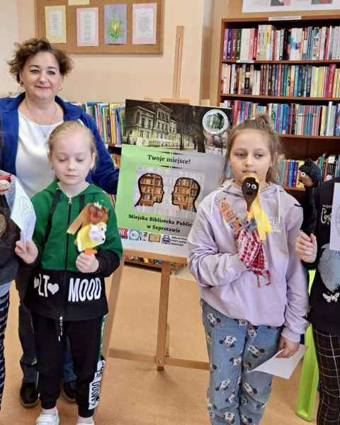 zdjęcie bibliotekarki i dzieci z kukiełkami przy plakacie biblioteki