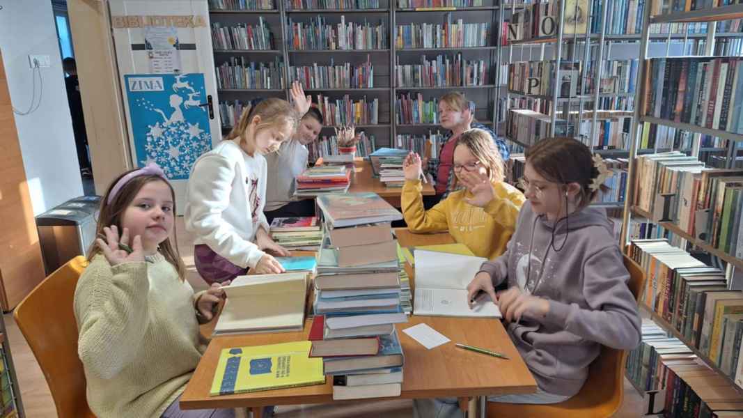 zdjęcie dzieci przy stolikach z książkami w bibliotece machają rękami, podczas ubytkowania książek