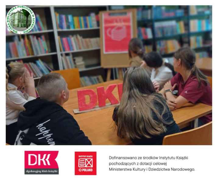 zdjęcie dzieci przy stoliku patrzących na plakat DKK, po lewej logo biblioteki, na dole belka z logo DKK, instytutu książki oraz informacja o dofinansowaniu ze środków Instytutu książki