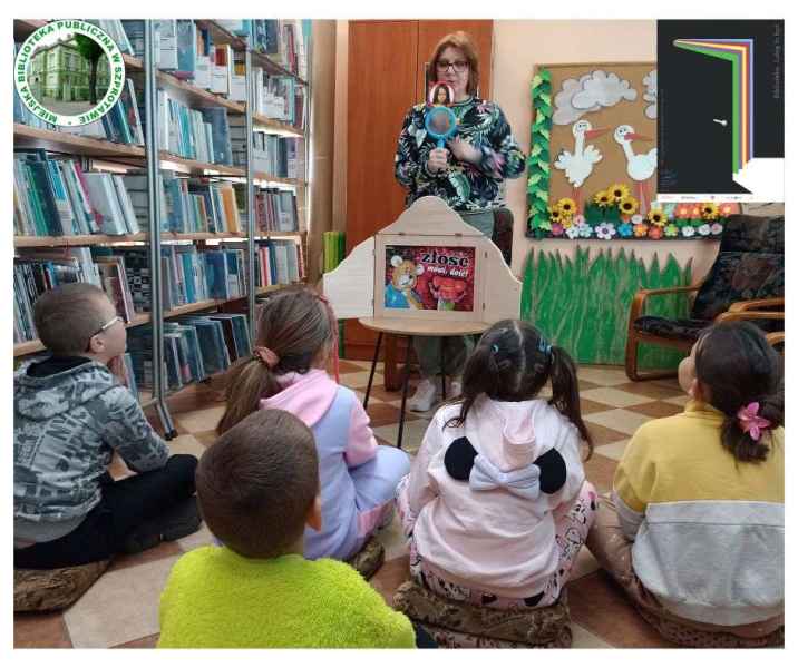 zdjęcie bibliotekarki pokazującej dzieciom lusterko emocji, po lewej logo biblioteki