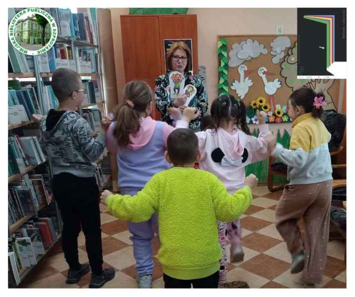 zdjęcie bibliotekarki i dzieci z ilustracjami do teatrzyku kamishibai, po lewej logo biblioteki