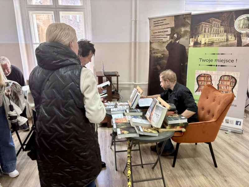 zdjęcie autora podczas rozdawania autografów czytelnikom przy stoliku z książkami 