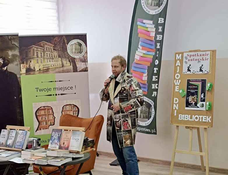 zdjęcie autora w płaszczu z okładek książek podczas mówienia do widowni, obok stolik z książkami, w tle flaga i baner biblioteki