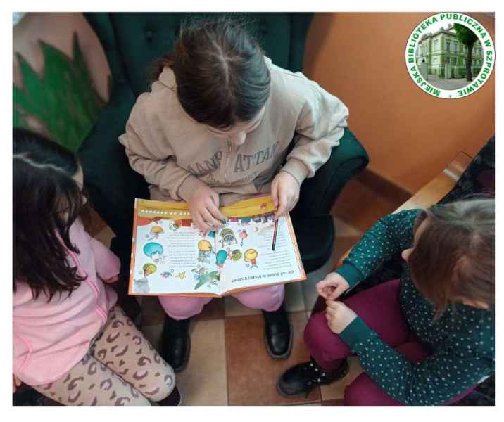 zdjęcie dziewczynek siedzących w fotelach z książką, po prawej logo biblioteki