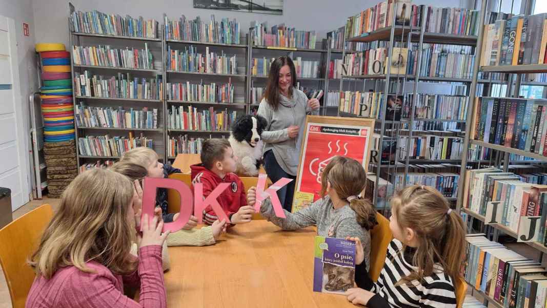 zdjęcie dzieci przy stoliku z papierowym napisem DKK i książką, z tyłu nauczycielka, maskotka psa i plakat DKK