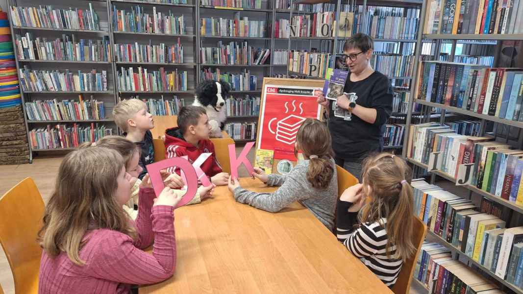 zdjęcie dzieci przy stoliku z papierowym napisem DKK, bibliotekarką pokazującą książkę, z tyłu maskotka psa i plakat DKK