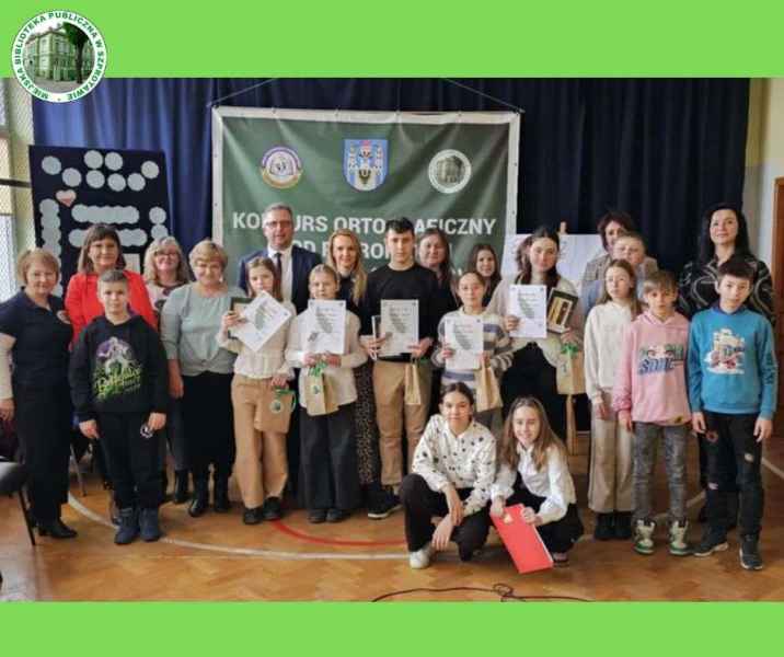 zdjęcie uczestników i zwycięzców konkursu z nagrodami w gronie burmistrza, nauczycielek, bibliotekarek i komisji konkursowej, po lewej logo biblioteki