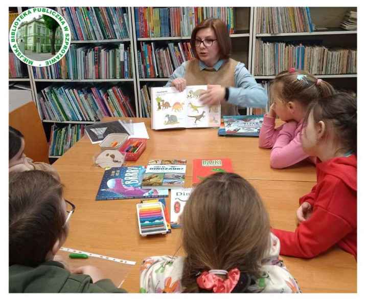 zdjęcie bibliotekarki przy stoliku z dziećmi pokazującej książkę z dinozaurami, po lewej logo biblioteki