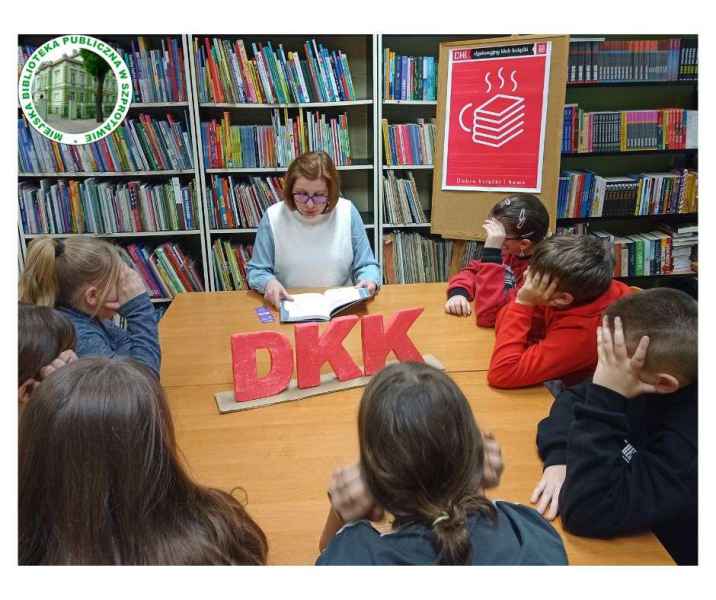 zdjęcie czytającej bibliotekarki i dzieci przy stoliku ze styropianowym napisem DKK, w tle plakat DKK, po lewej logo biblioteki